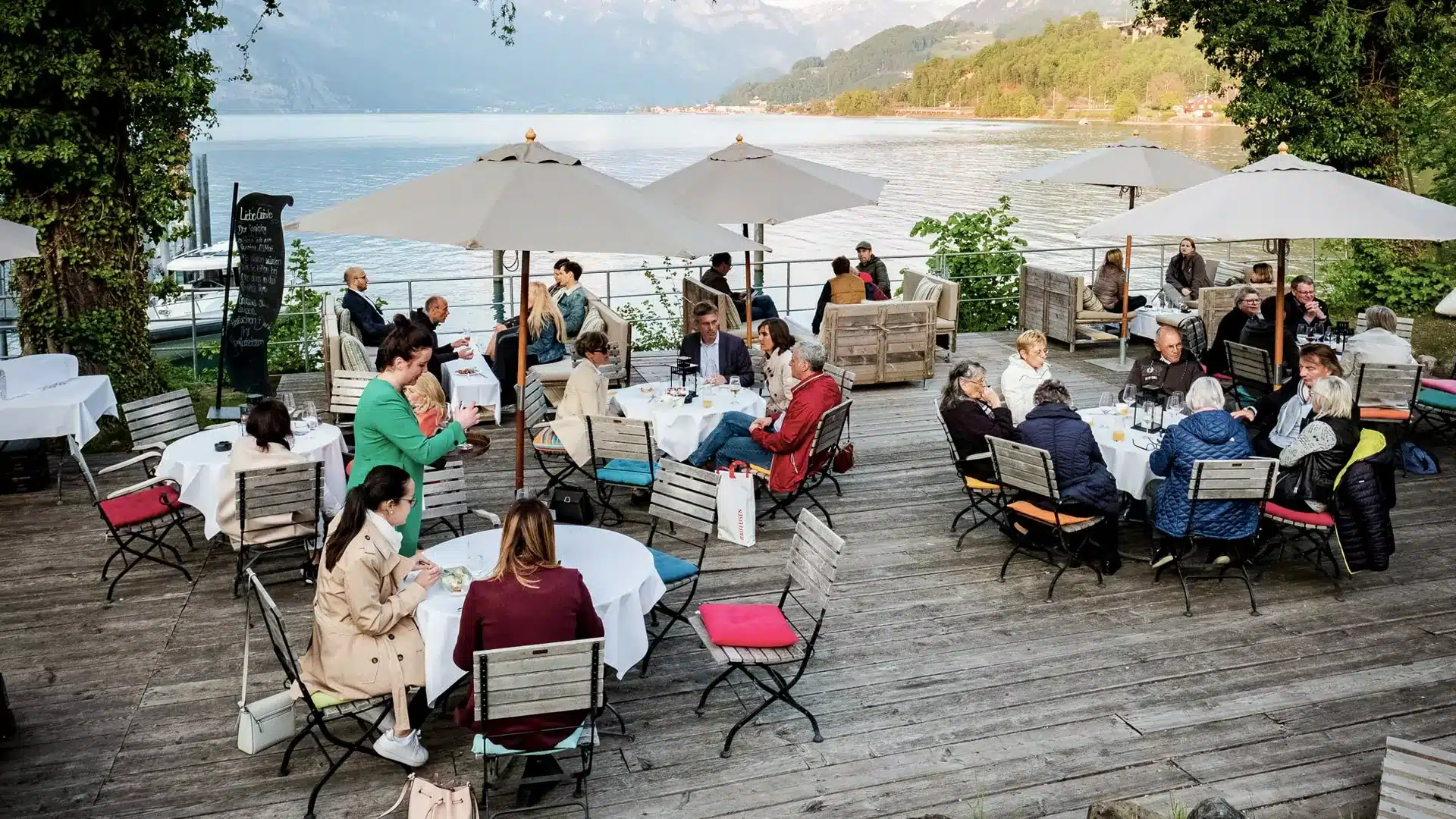 Menschengruppen sitzen auf der Plattform am See in der Sagibeiz unter Sonnenschirmen.